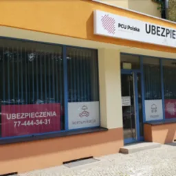 Fasada budynku agencji ubezpieczeniowej PCU Polska z widocznymi reklamami okiennymi dotyczącymi ubezpieczeń majątkowych, komunikacyjnych i na życie.
