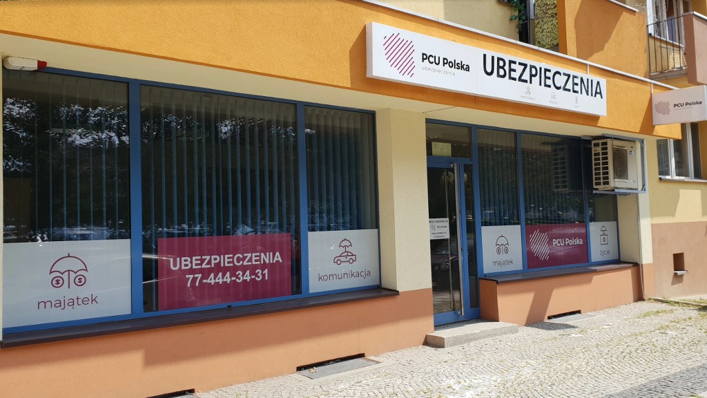 Fasada budynku agencji ubezpieczeniowej PCU Polska z widocznymi reklamami okiennymi dotyczącymi ubezpieczeń majątkowych, komunikacyjnych i na życie.