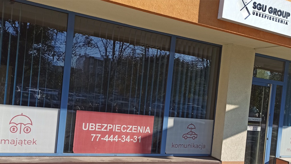 Fasada budynku z witryną agencji ubezpieczeniowej SGU Group, na szybach reklamy dotyczące ubezpieczeń majątku i komunikacji z numerem telefonu, w tle zaparkowane samochody.