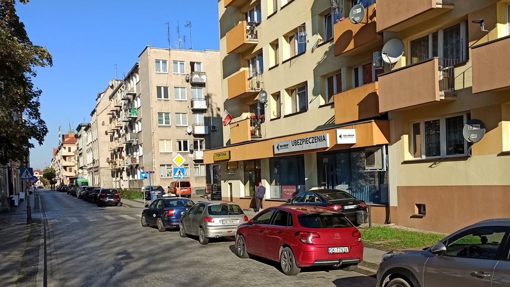 Ulica miejska z zaparkowanymi samochodami, w tle budynek z szyldem 'Ubezpieczenia'