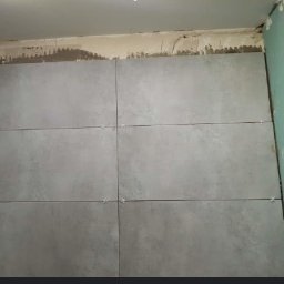 Marcin Rosiak - Ściana z szarych płytek ceramicznych w trakcie układania, krzyżyki dystansowe w spoinach, widoczny fragment ściany z surowym tynkiem i płyta gipsowo-kartonowa.