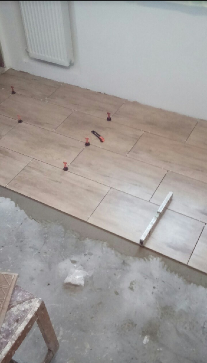 Układanie podłogi z płytek imitujących drewno z użyciem poziomicy i klipsów poziomujących, widoczny fragment starej podłogi i grzejnik na ścianie.