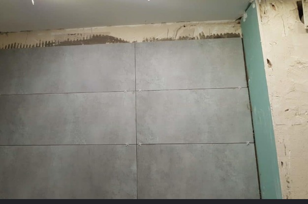 Ściana z szarych płytek ceramicznych w trakcie układania, krzyżyki dystansowe w spoinach, widoczny fragment ściany z surowym tynkiem i płyta gipsowo-kartonowa.