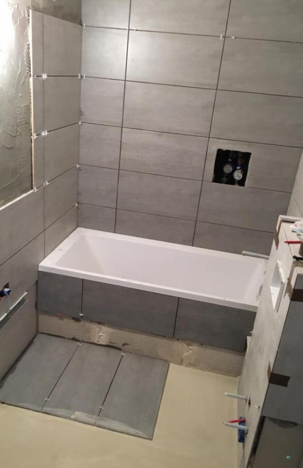 Układanie szarych płytek w łazience, widoczna wanna, płytki na ścianach z krzyżykami dystansowymi, przygotowane płytki na podłodze, instalacja wodna w ścianie.