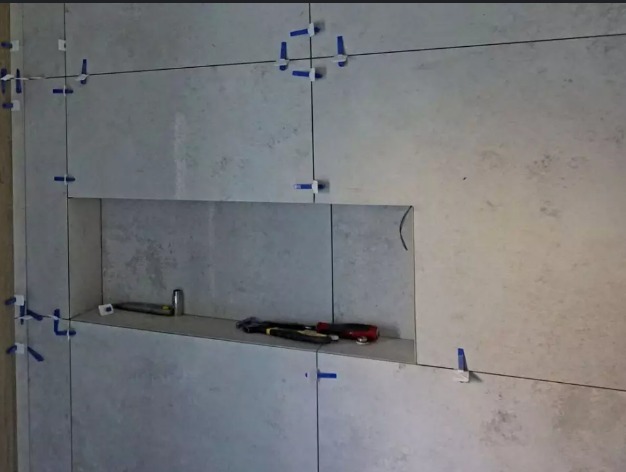 Ściana w trakcie układania szarych płytek z wnęką, widoczne niebieskie krzyżyki dystansowe, poziomica i narzędzia glazurnicze.