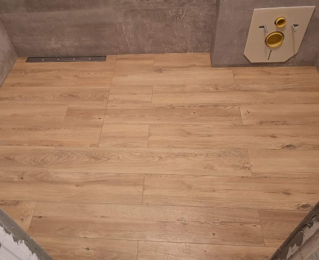 Ułożona podłoga z płytek imitujących drewno w łazience, widoczny odpływ liniowy przy ścianie i zabudowa stelaża podtynkowego WC.