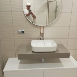 Zakład Remontowo Budowlany Cojg Zbigniew - Nowoczesna umywalka nablatowa w łazience z beżowymi płytkami, okrągłe lustro z widocznym odbiciem osoby robiącej zdjęcie. Minimalistyczny design i czyste linie.