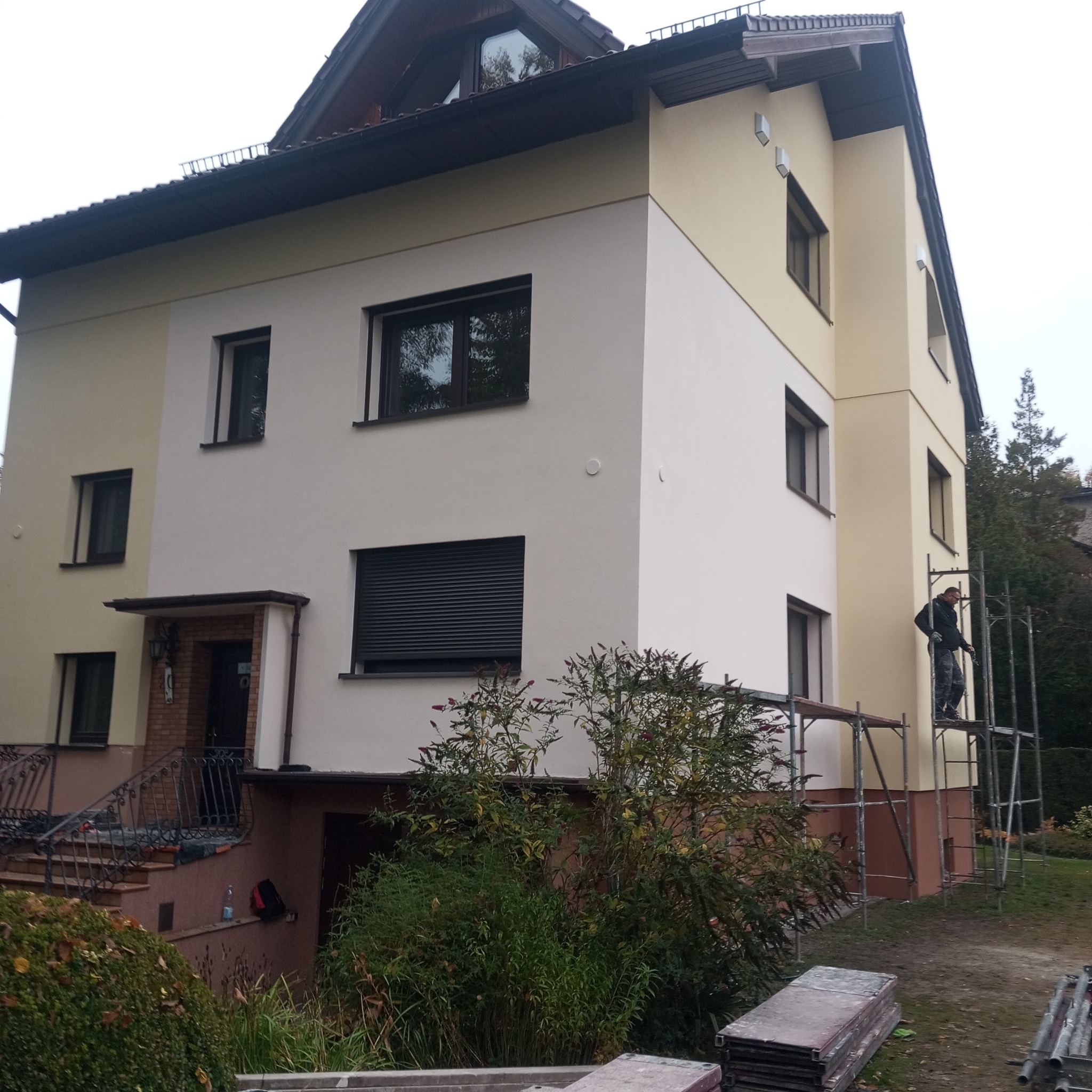 Dwukondygnacyjny dom z ocieploną elewacją w odcieniach beżu i bieli. Pracownik na rusztowaniu maluje ścianę. Widoczne okna z roletami i schody wejściowe.