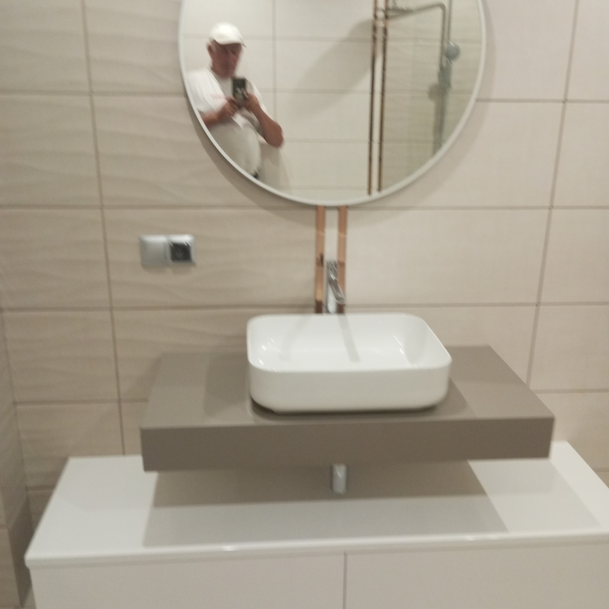 Nowoczesna umywalka nablatowa w łazience z beżowymi płytkami, okrągłe lustro z widocznym odbiciem osoby robiącej zdjęcie. Minimalistyczny design i czyste linie.