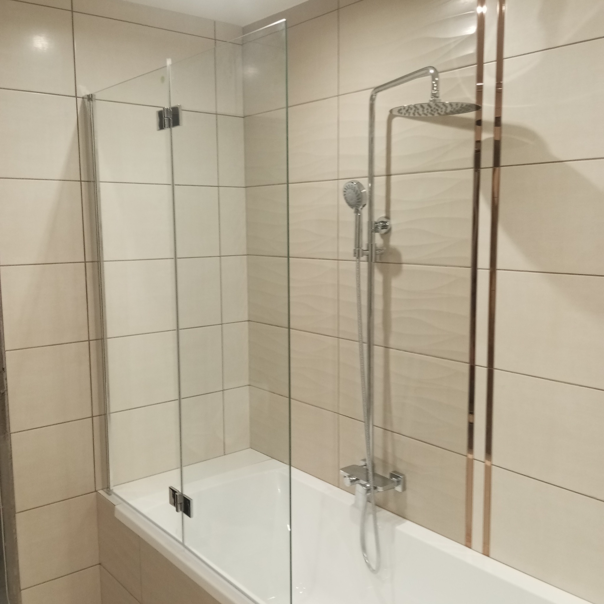 Beżowa łazienka po remoncie: kabina prysznicowa ze szklanym parawanem, bateria i deszczownica. W tle płytki z delikatnym wzorem i dekoracyjnymi, złotymi paskami.