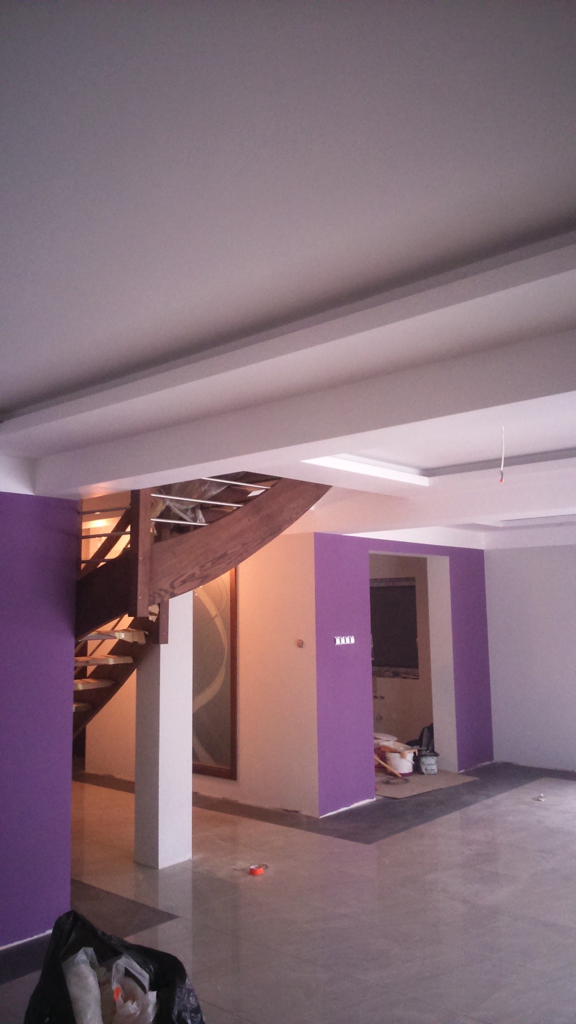 Wnętrze w trakcie malowania: fioletowe akcenty na ścianach, drewniane schody i sufit z oświetleniem. Widoczne ślady prac remontowych, w tym worki i narzędzia.