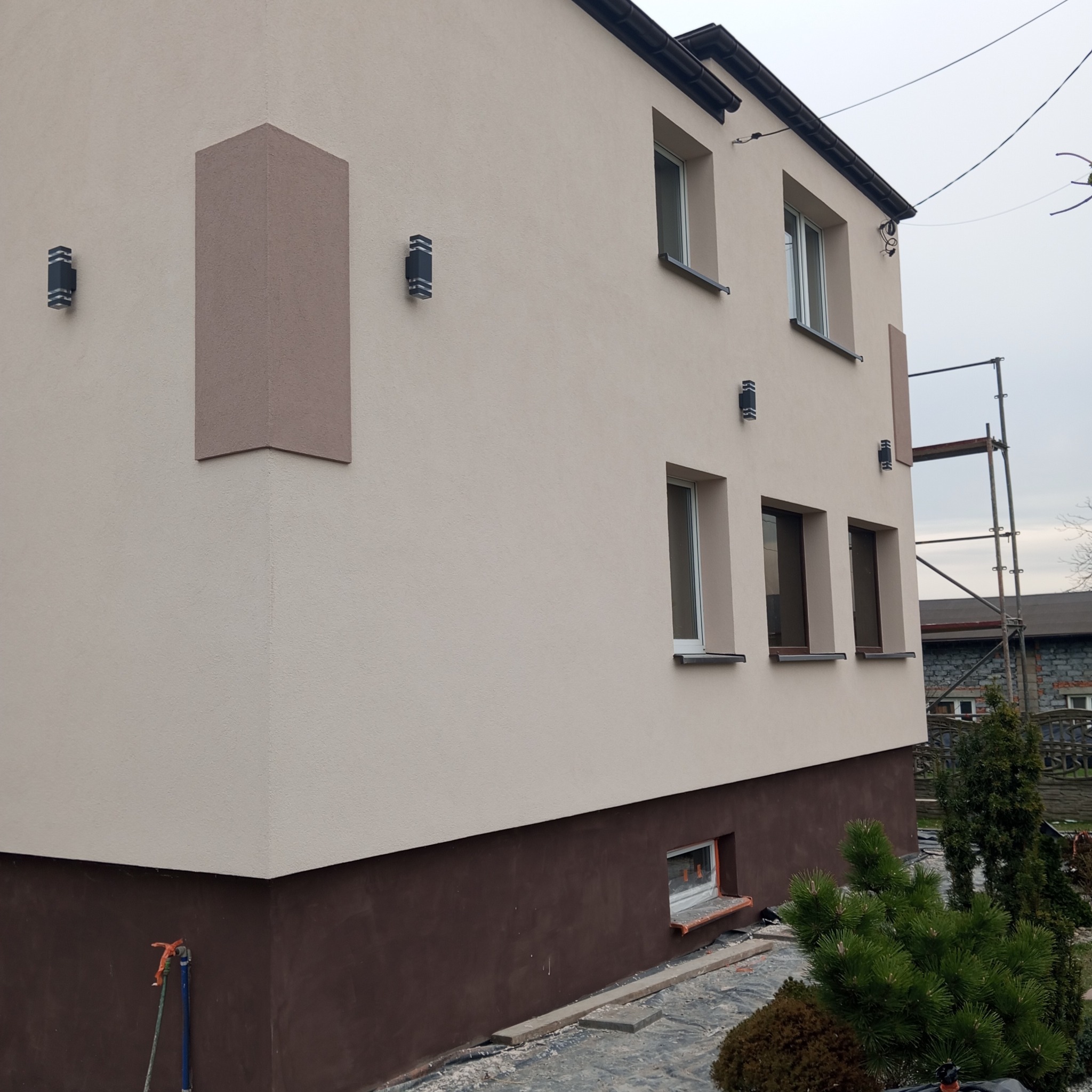Dwukolorowa elewacja domu po remoncie z dekoracyjnymi elementami. Widoczne nowe lampy, okna. Częściowo widoczne rusztowanie.