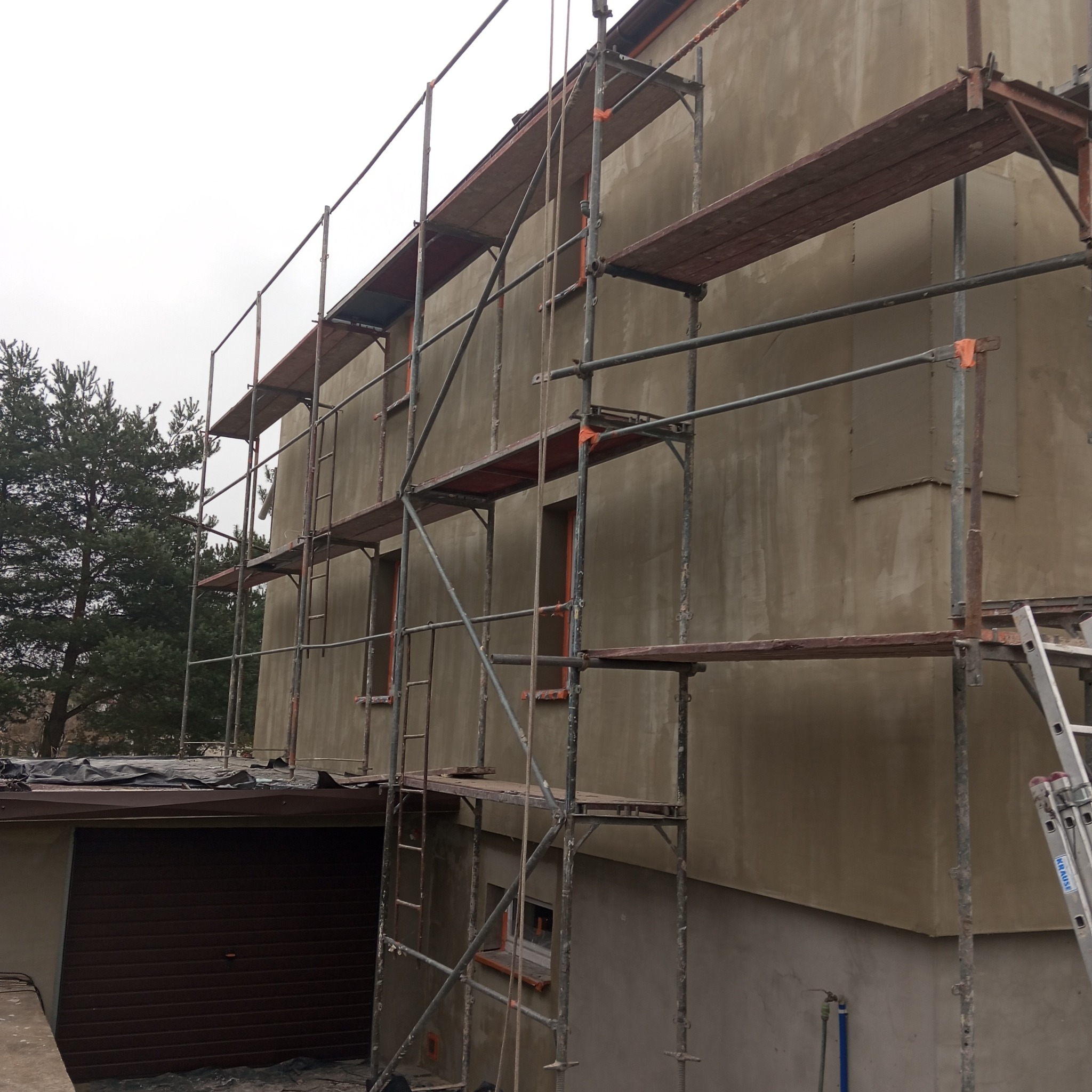 Budynek w trakcie prac elewacyjnych, widoczne rusztowania i świeżo nałożony tynk w odcieniach beżu. Częściowo widoczny garaż z brązową bramą i zieleń drzew w tle.