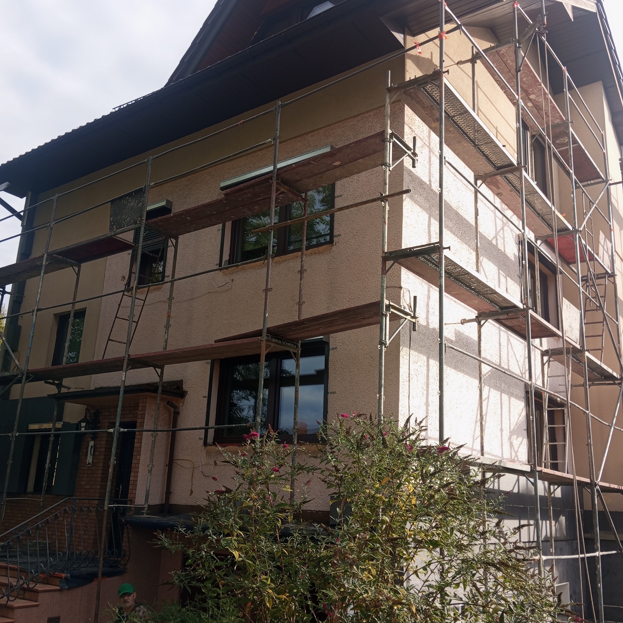 Dom w rusztowaniach, elewacja w trakcie prac. Widoczne okna z ciemnymi ramami, krzew przed budynkiem. Ujęcie z dołu, częściowo pochmurne niebo.