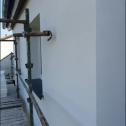 Świeżo pomalowana, jasna elewacja budynku z widocznymi rusztowaniami i oknem z czarnym parapetem; widoczna faktura tynku.