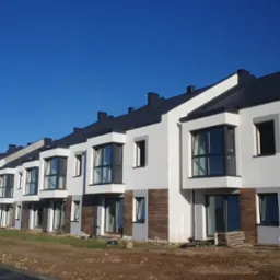 Szereg identycznych, nowo wybudowanych domów jednorodzinnych z białymi elewacjami, ciemną stolarką okienną i grafitowym dachem, sfotografowanych w słoneczny dzień na tle błękitnego nieba.