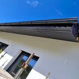 Wandmax - Biała elewacja domu z czarnym dachem i oknami, widziana z rusztowania. Świeżo pomalowana ściana, błękitne niebo w tle. Ujęcie z dołu, podkreślające detale wykończenia.