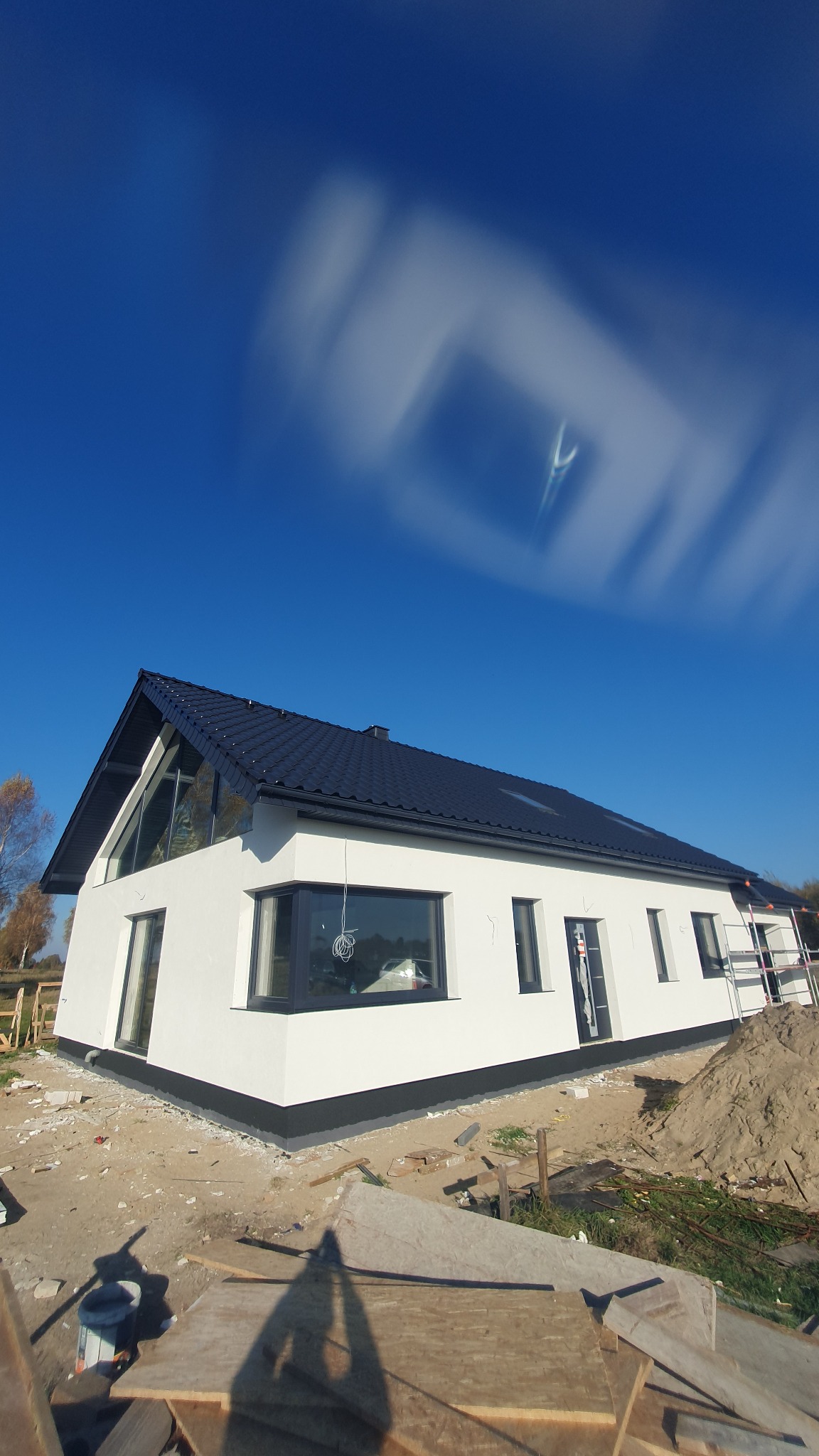 Nowoczesny dom jednorodzinny z białą elewacją i ciemną stolarką okienną oraz dachem, widoczny na tle błękitnego nieba z rozmyciem ruchu chmur, teren wokół domu w trakcie prac wykończeniowych.