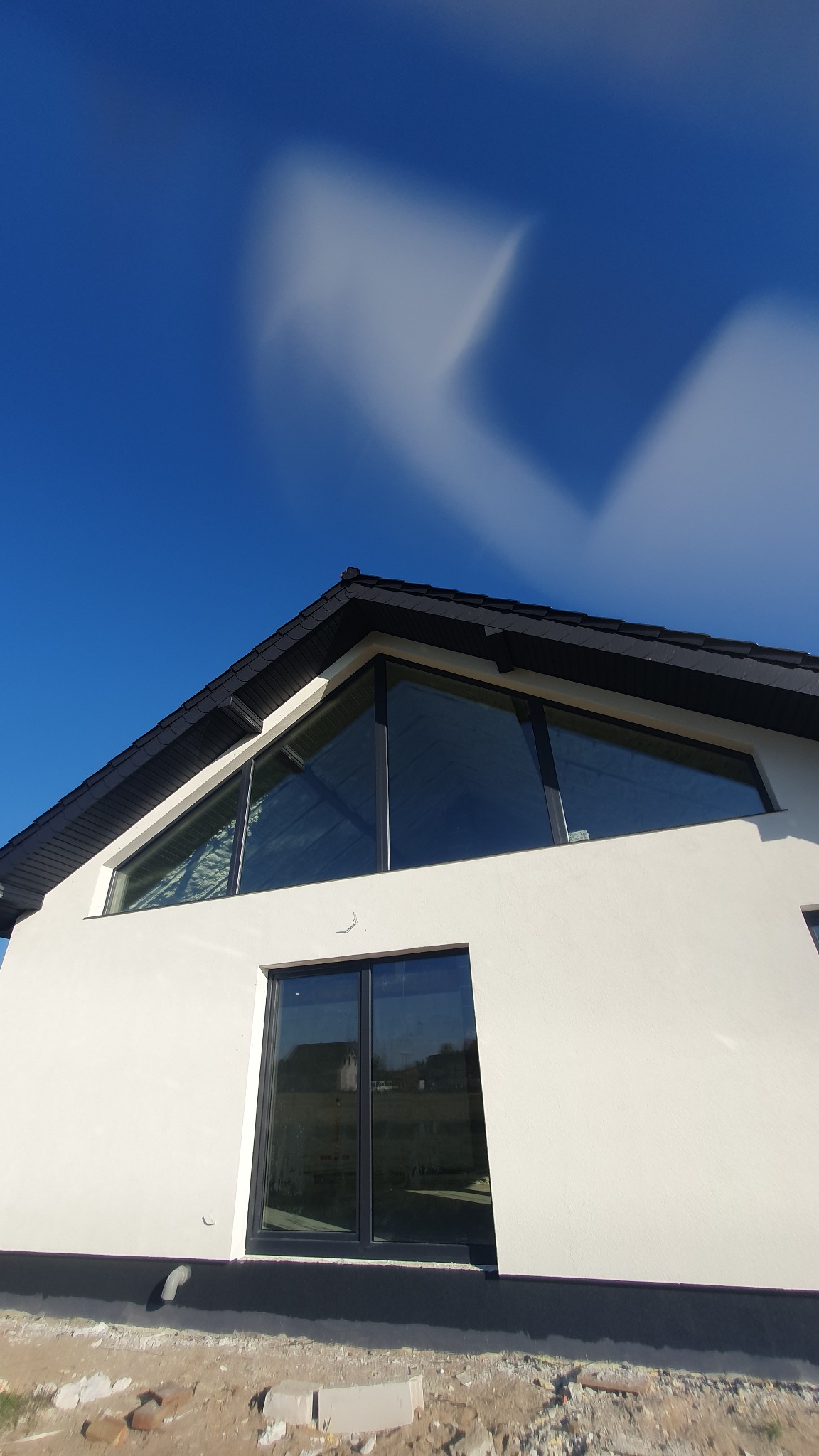 Nowoczesny dom z białymi ścianami, dużymi oknami z czarnymi ramami i czarnym dachem na tle błękitnego nieba z przesuwającymi się chmurami.