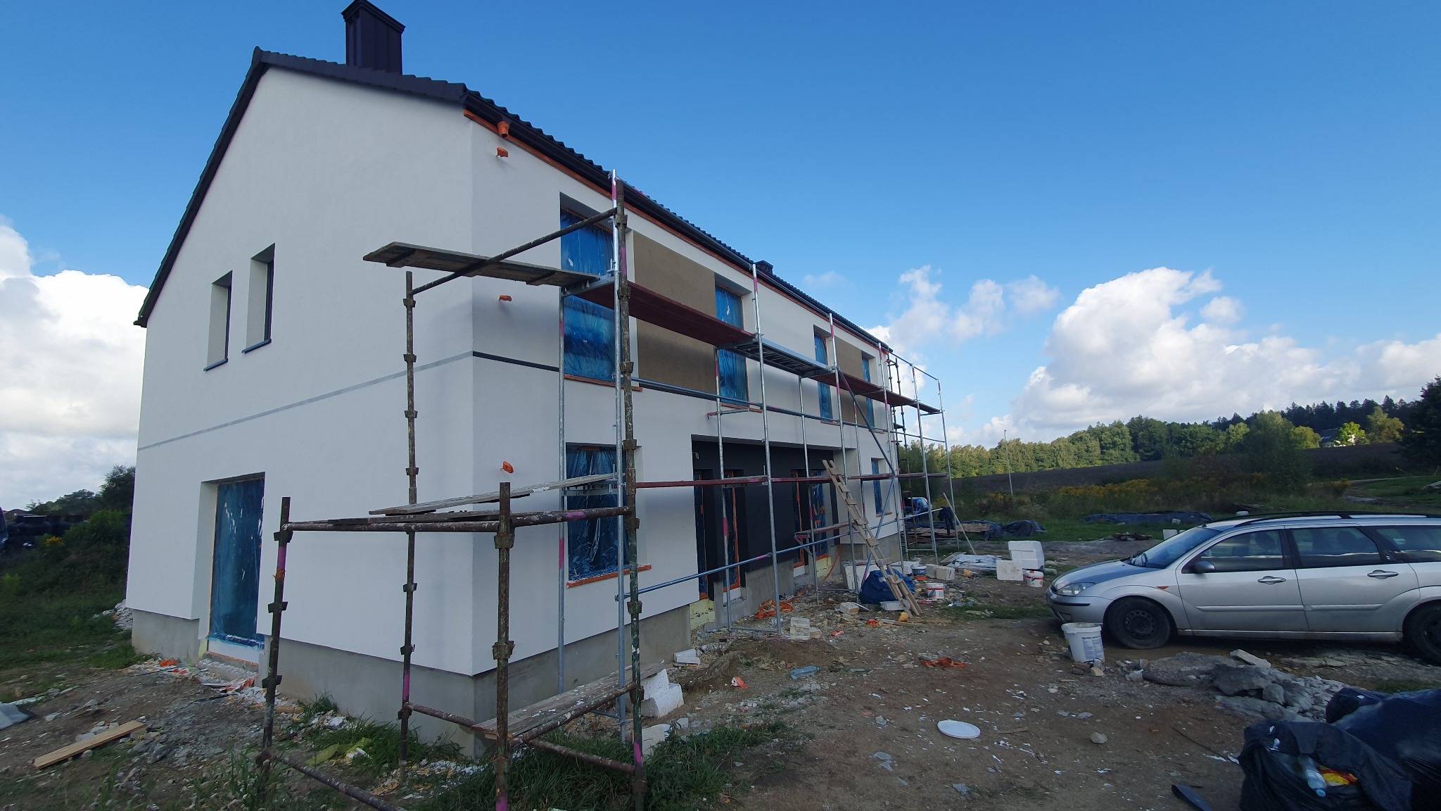 Dwukondygnacyjny dom w trakcie prac elewacyjnych, z oknami zabezpieczonymi folią, otoczony rusztowaniami, z widocznymi elementami krajobrazu i samochodem osobowym w tle.