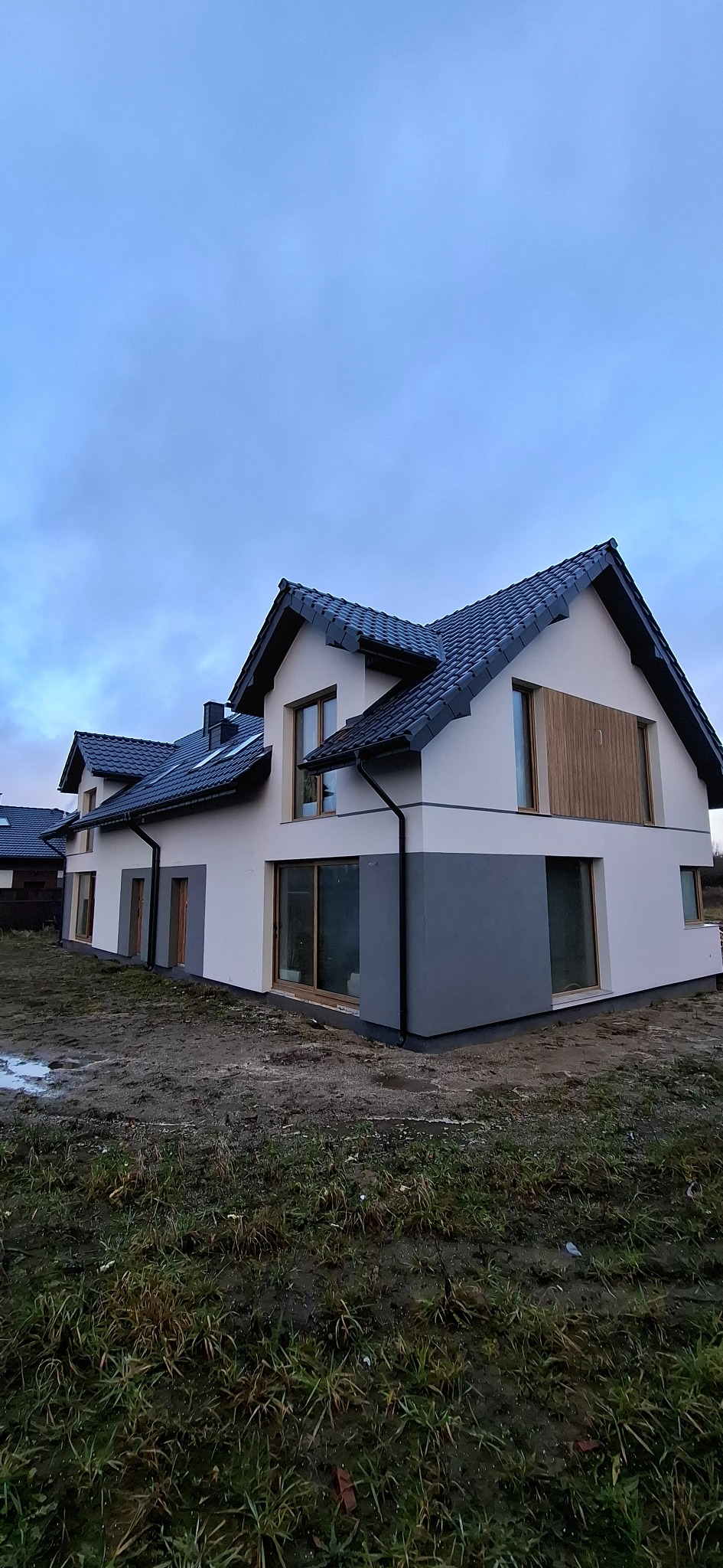 Dwukondygnacyjny dom z ciemną dachówką i jasną elewacją, z szarym pasem na dole. Widoczne okna i rynny. Trawa wokół domu jest błotnista. Pochmurne niebo w tle.