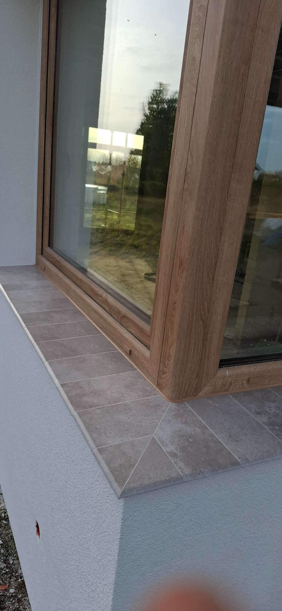 Szczegółowe ujęcie okna z drewnianą ramą i szarym parapetem z płytek, osadzonym w jasnoszarej elewacji budynku. Widoczne odbicie otoczenia w szybie.