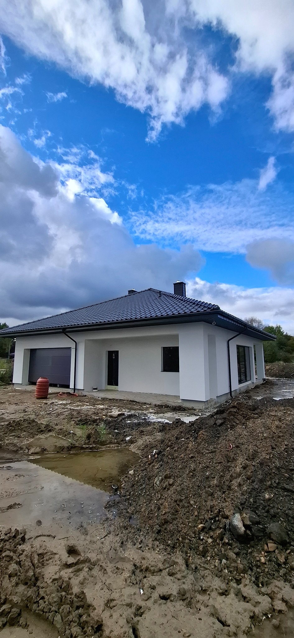 Nowoczesny, parterowy dom z białymi ścianami i ciemną dachówką w Słupsku. Widoczne okna, brama garażowa i fragment terenu przed budynkiem.