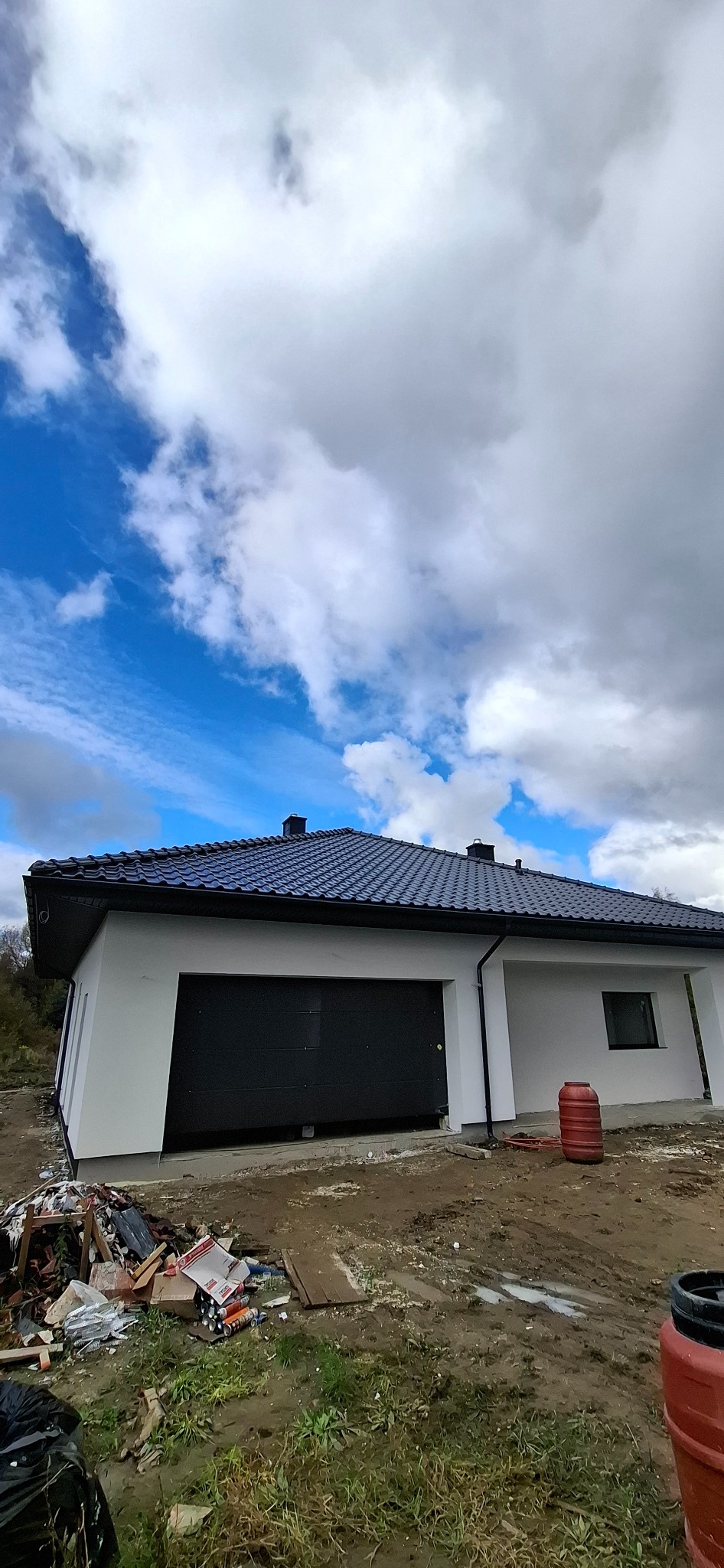 Nowoczesny dom z garażem i ciemną dachówką na tle pochmurnego nieba. Biała elewacja, widoczne detale wykończeniowe. Teren wokół domu w trakcie prac.