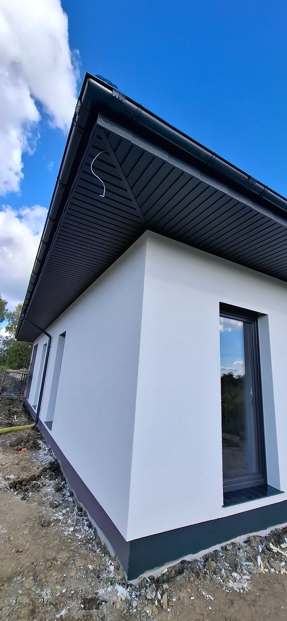 Biała elewacja domu jednorodzinnego z ciemną podbitką i oknami, widok z dołu w słoneczny dzień. Teren wokół domu jeszcze niezagospodarowany.