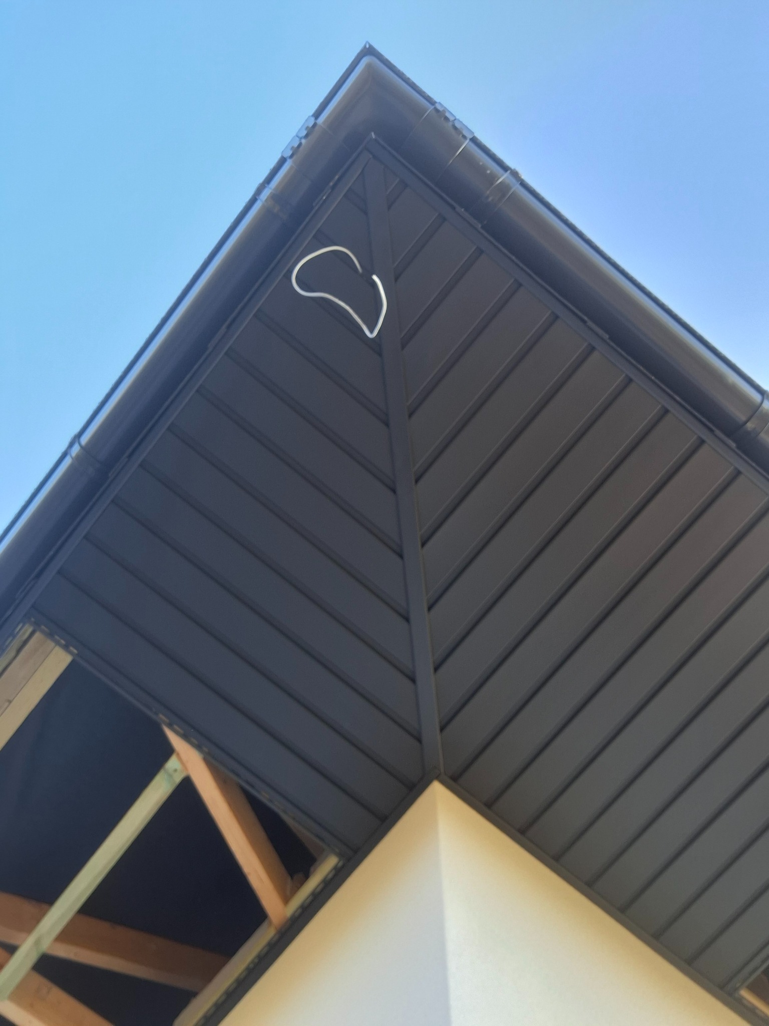 Szare podbitki dachowe z widocznym fragmentem konstrukcji drewnianej i orynnowaniem na tle błękitnego nieba, ujęcie z dołu.