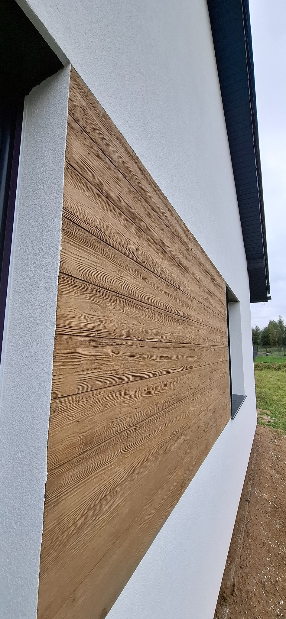 Fragment elewacji domu z dekoracyjnym panelem imitującym drewno, widocznym z boku. Biały tynk kontrastuje z brązowym panelem i czarną ramą okienną.