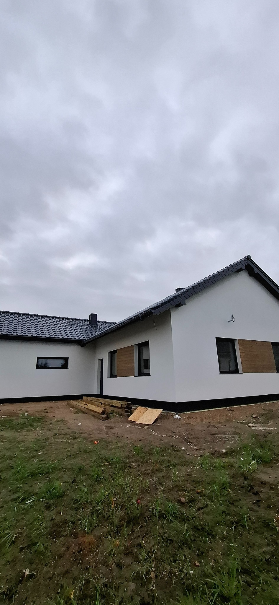 Biały dom z ciemną dachówką i czarnymi ramami okiennymi. Część okien zabita deskami. Na ziemi leżą deski. Pochmurne niebo.