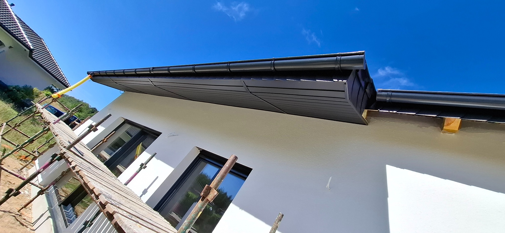 Biała elewacja domu z czarnym dachem i oknami, widziana z rusztowania. Świeżo pomalowana ściana, błękitne niebo w tle. Ujęcie z dołu, podkreślające detale wykończenia.