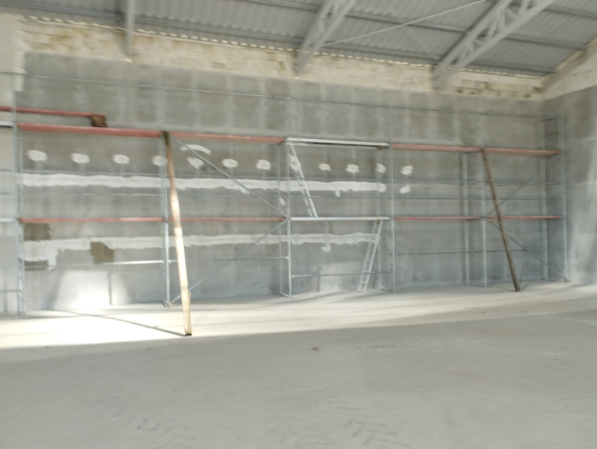 Wnętrze hali w trakcie prac wykończeniowych, widoczne rusztowania przy ścianie z częściowo nałożoną warstwą szpachli, surowy betonowy strop.