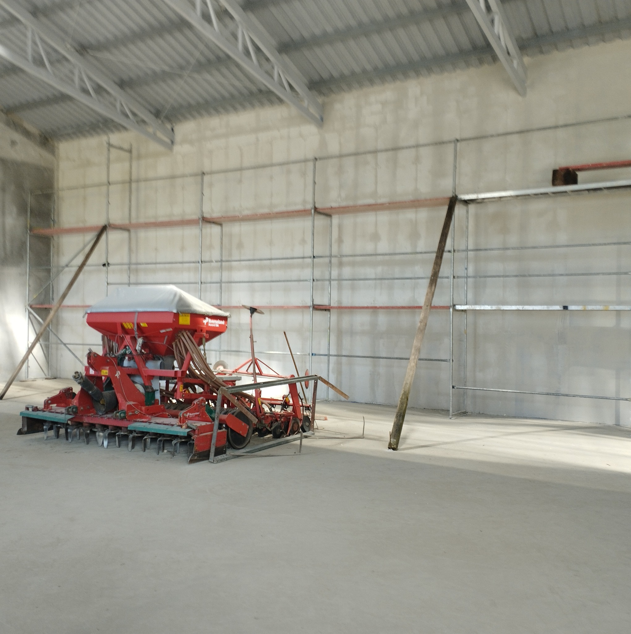 Wnętrze hali z betonową posadzką i rusztowaniami przy ścianach, w centrum czerwony siewnik rolniczy z szarą plandeką.