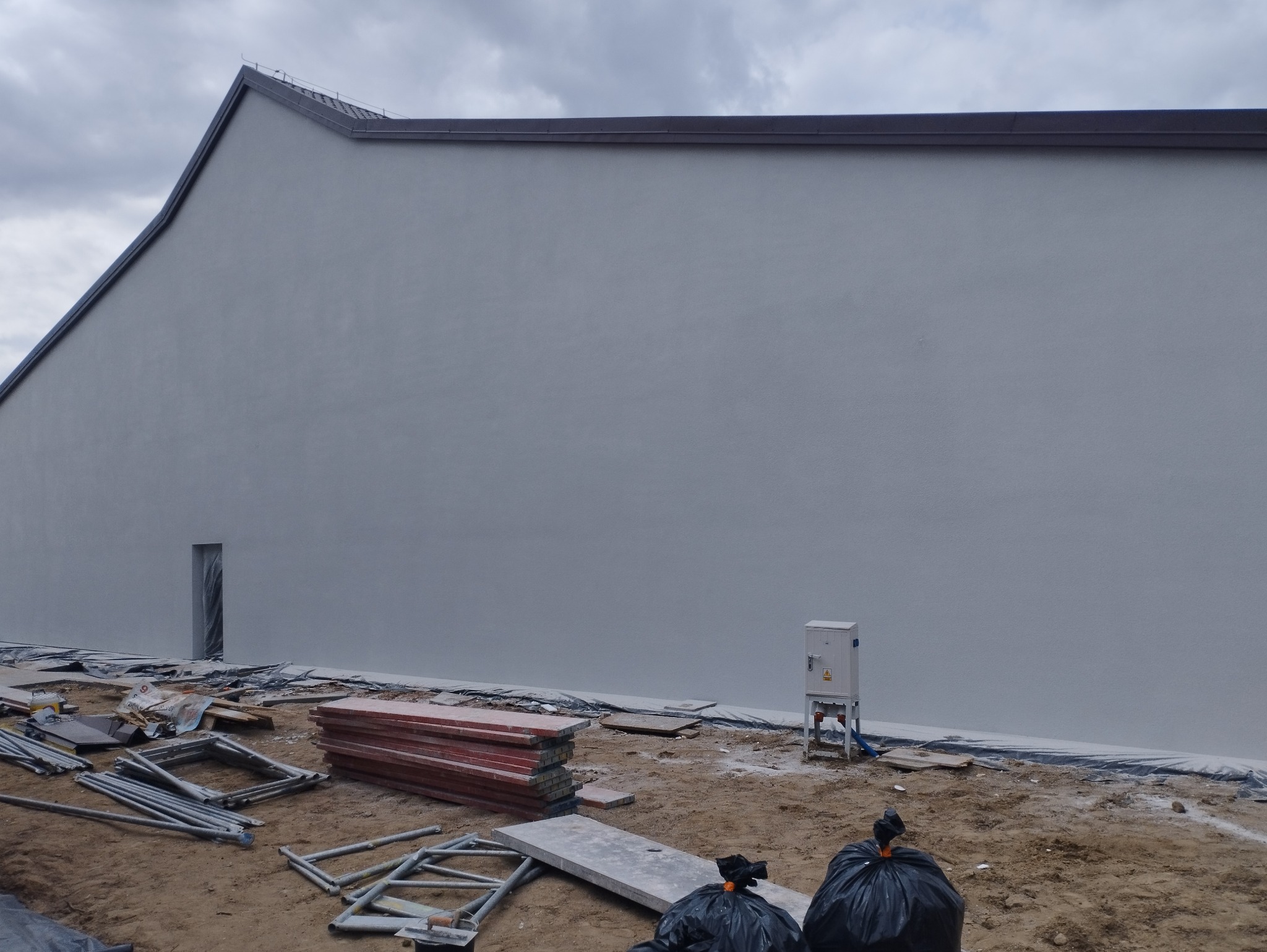 Świeżo otynkowana elewacja budynku z widocznymi śladami prac, rusztowaniami i skrzynką elektryczną na zewnątrz.