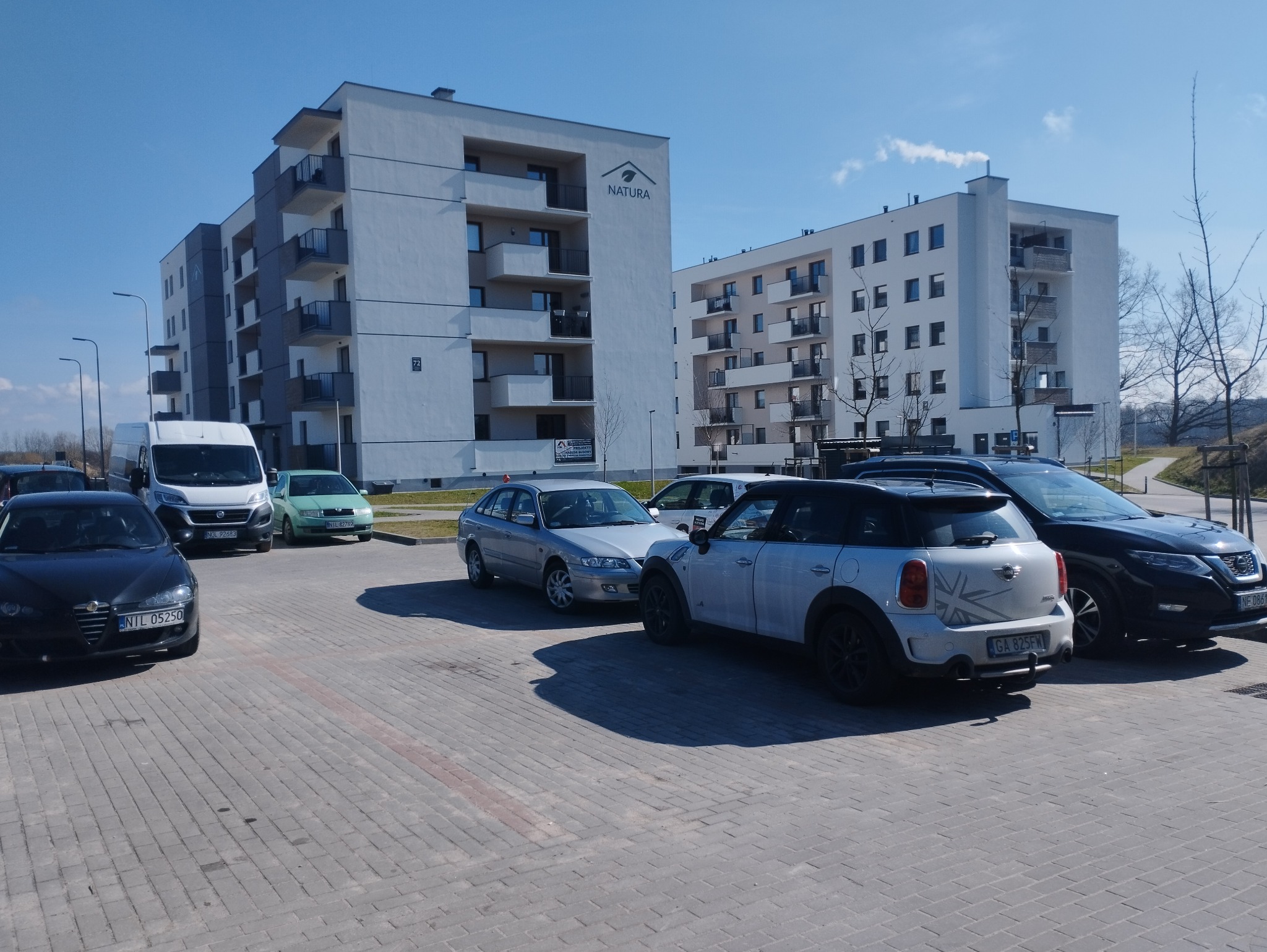 Nowoczesny, biały budynek wielorodzinny z balkonami, oświetlony jasnym słońcem, z logo dewelopera 'NATURA' widocznym na elewacji, otoczony zaparkowanymi samochodami na brukowanym parkingu.