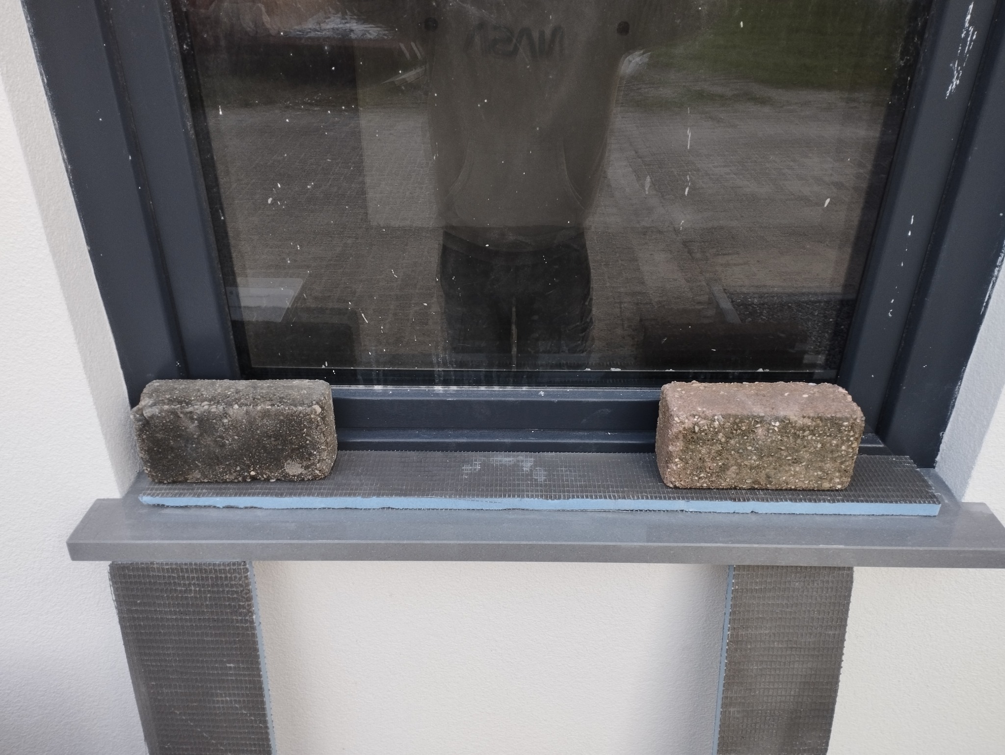 Przygotowanie do ocieplenia elewacji okna: styropian z siatką, podkładki z cegieł, rama okienna z widocznym odbiciem osoby.