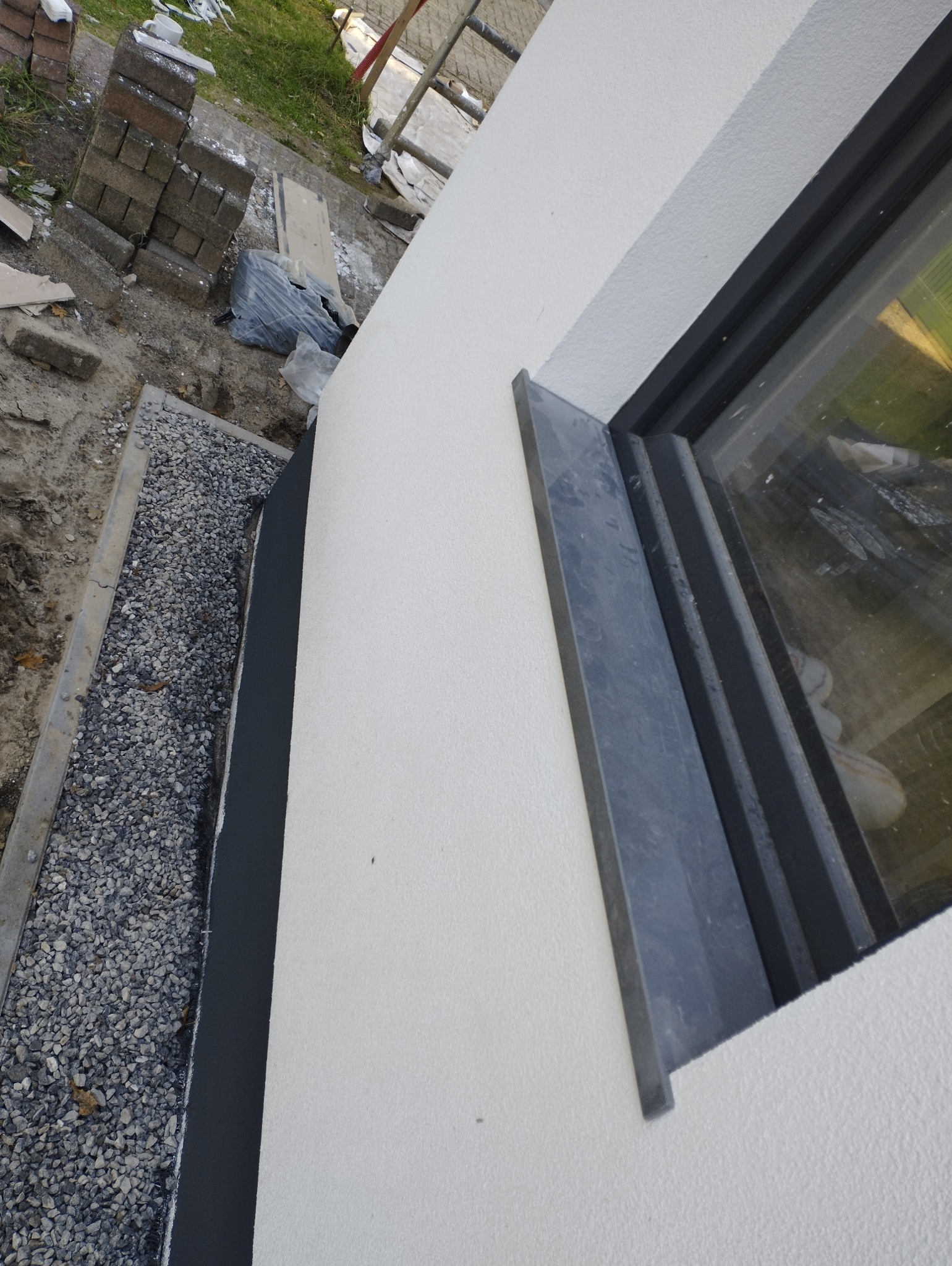 Fragment ściany budynku z oknem po nałożeniu jasnego tynku strukturalnego, widoczna obróbka okienna w kolorze grafitowym oraz przygotowany teren wokół budynku z podsypką z grysu.