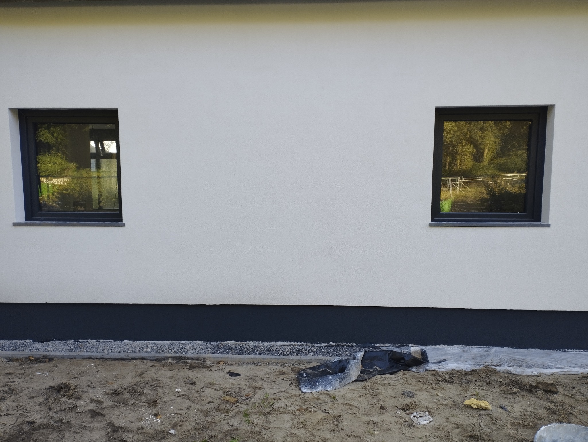 Ocieplona elewacja budynku z dwoma oknami, widoczne zabezpieczenia folią i ziemia przy fundamentach.