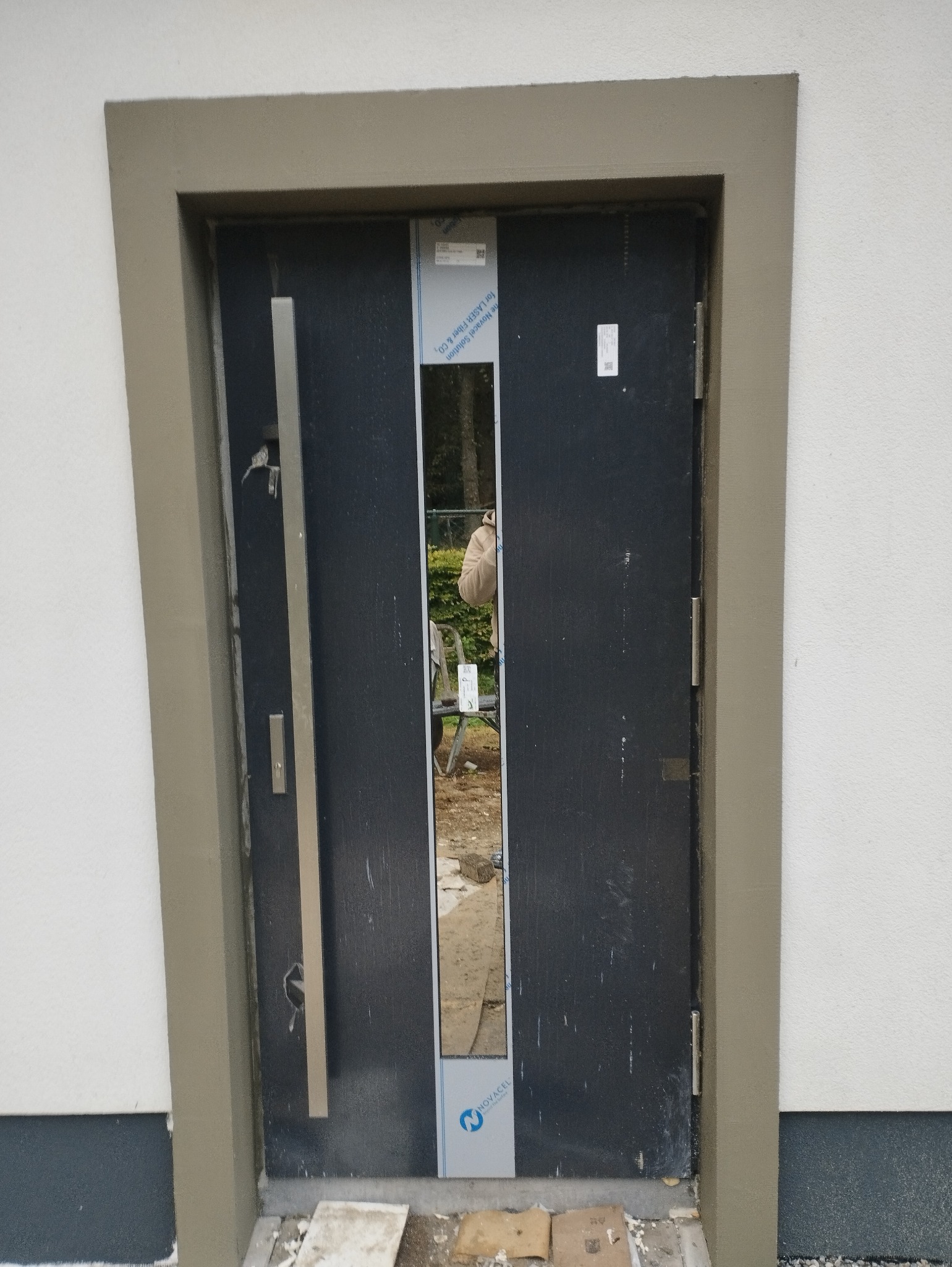 Nowe, czarne drzwi wejściowe z długim, pionowym uchwytem, osadzone w ościeżnicy w kolorze beżowym, w trakcie montażu na tle białej elewacji.