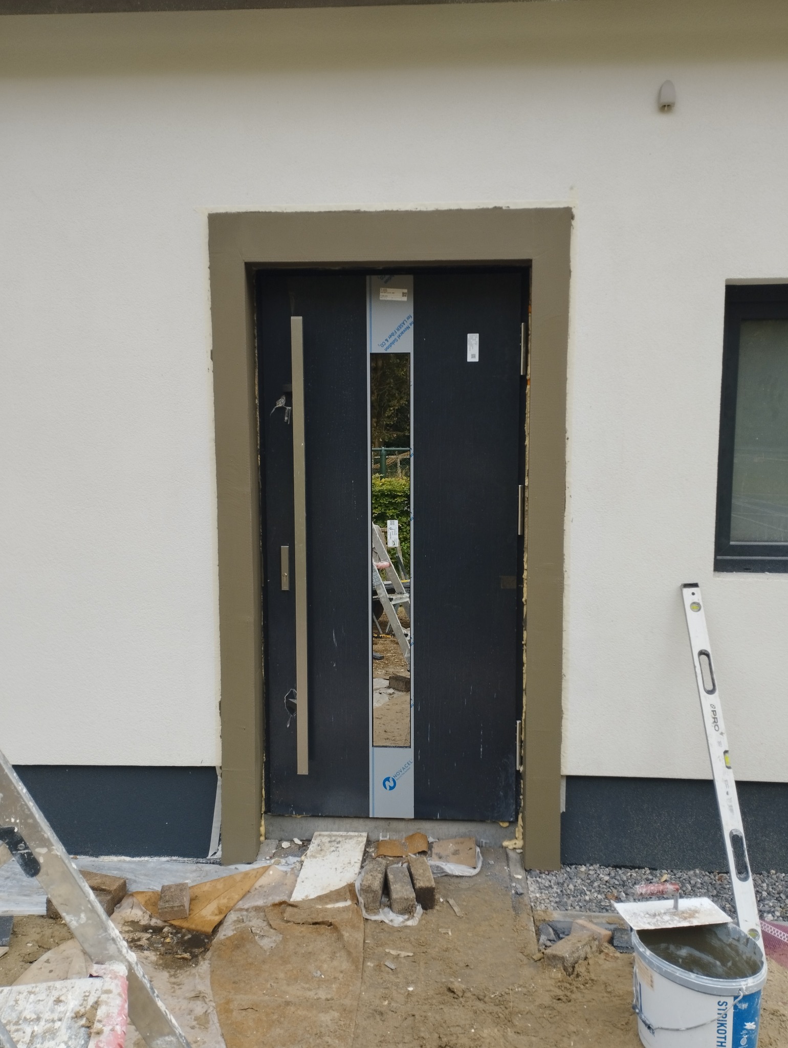 Wykończenie elewacji wokół czarnych drzwi wejściowych, widoczne świeżo nałożone obramowanie z zaprawy, poziomica oparta o ścianę, teren budowy z cegłami i wiadrem z zaprawą.