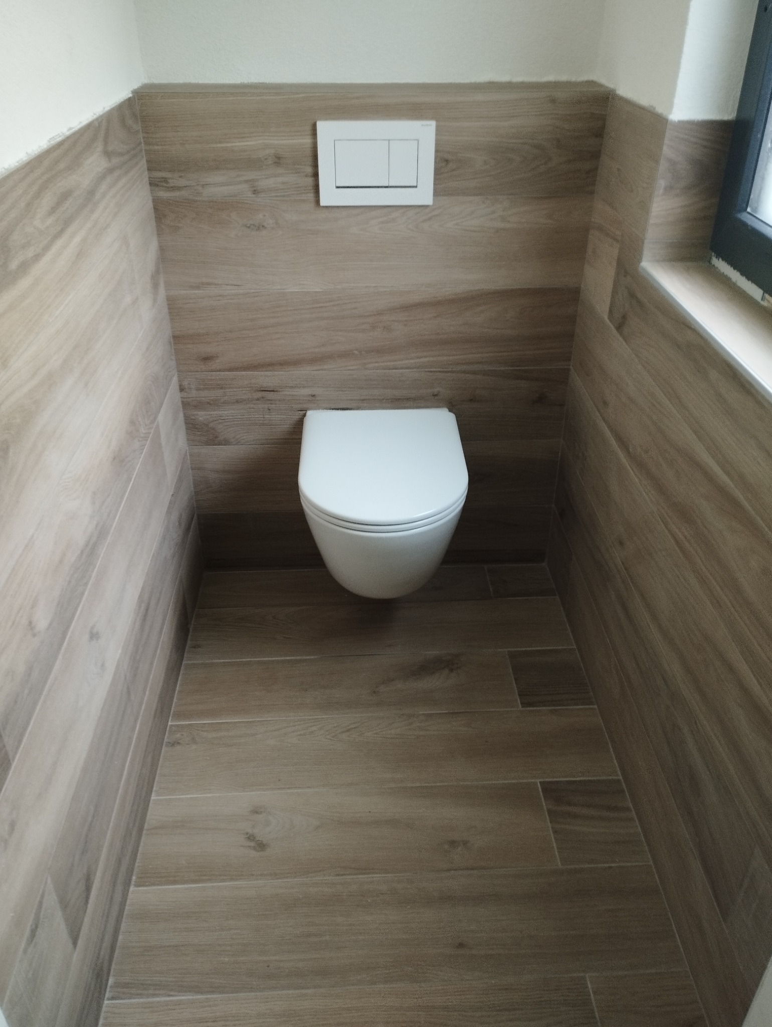 Zakończona aranżacja małej łazienki z wiszącą toaletą, wykończonej płytkami imitującymi drewno na podłodze i ścianach, z widocznym przyciskiem spłuczki.