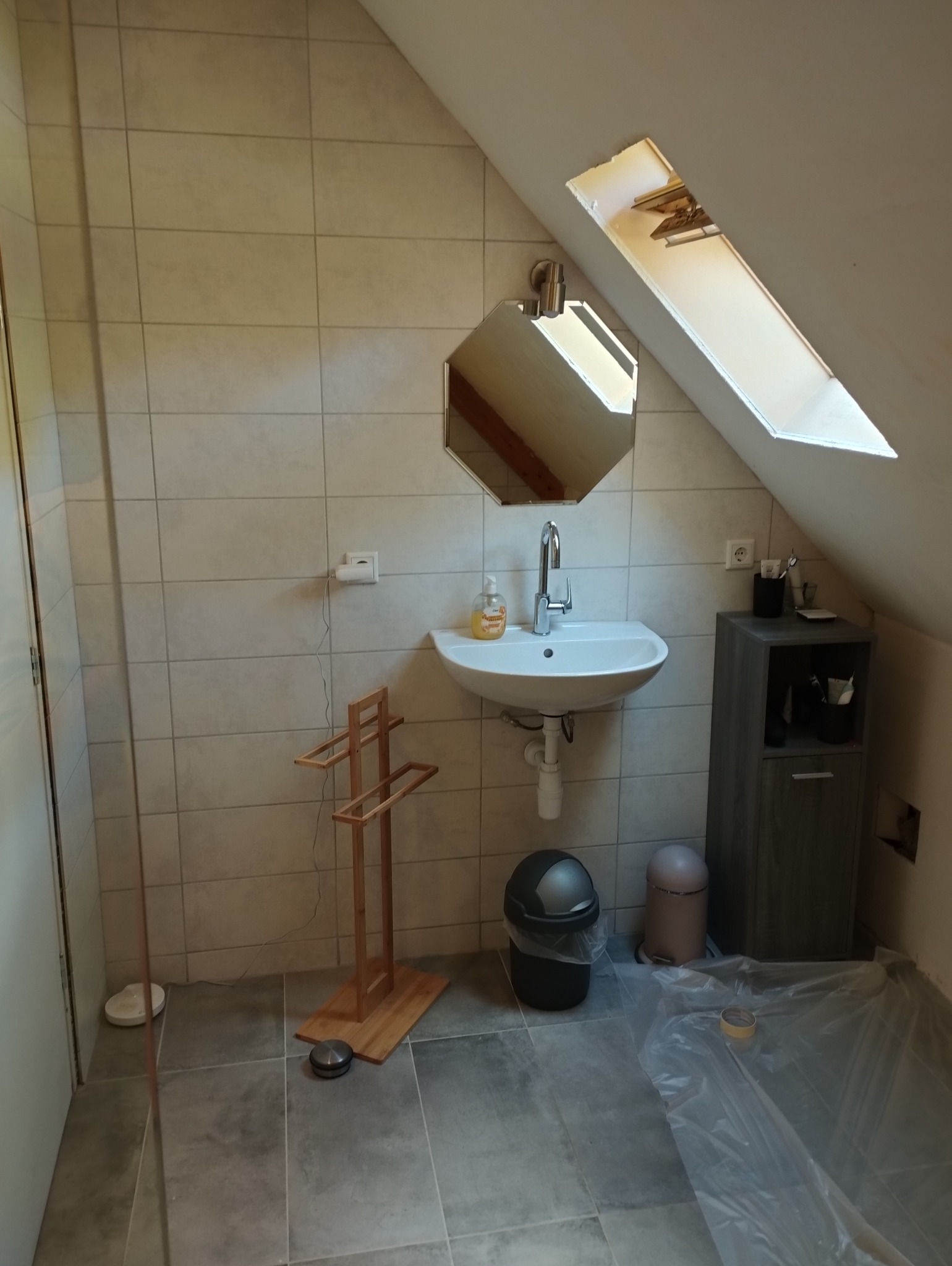 Umywalka z baterią i lustrem w ośmiokątnej ramie w łazience na poddaszu, widoczne płytki na ścianach i podłodze, zabezpieczona folią podłoga, drewniany wieszak na ręczniki i szafka z akcesoriami.