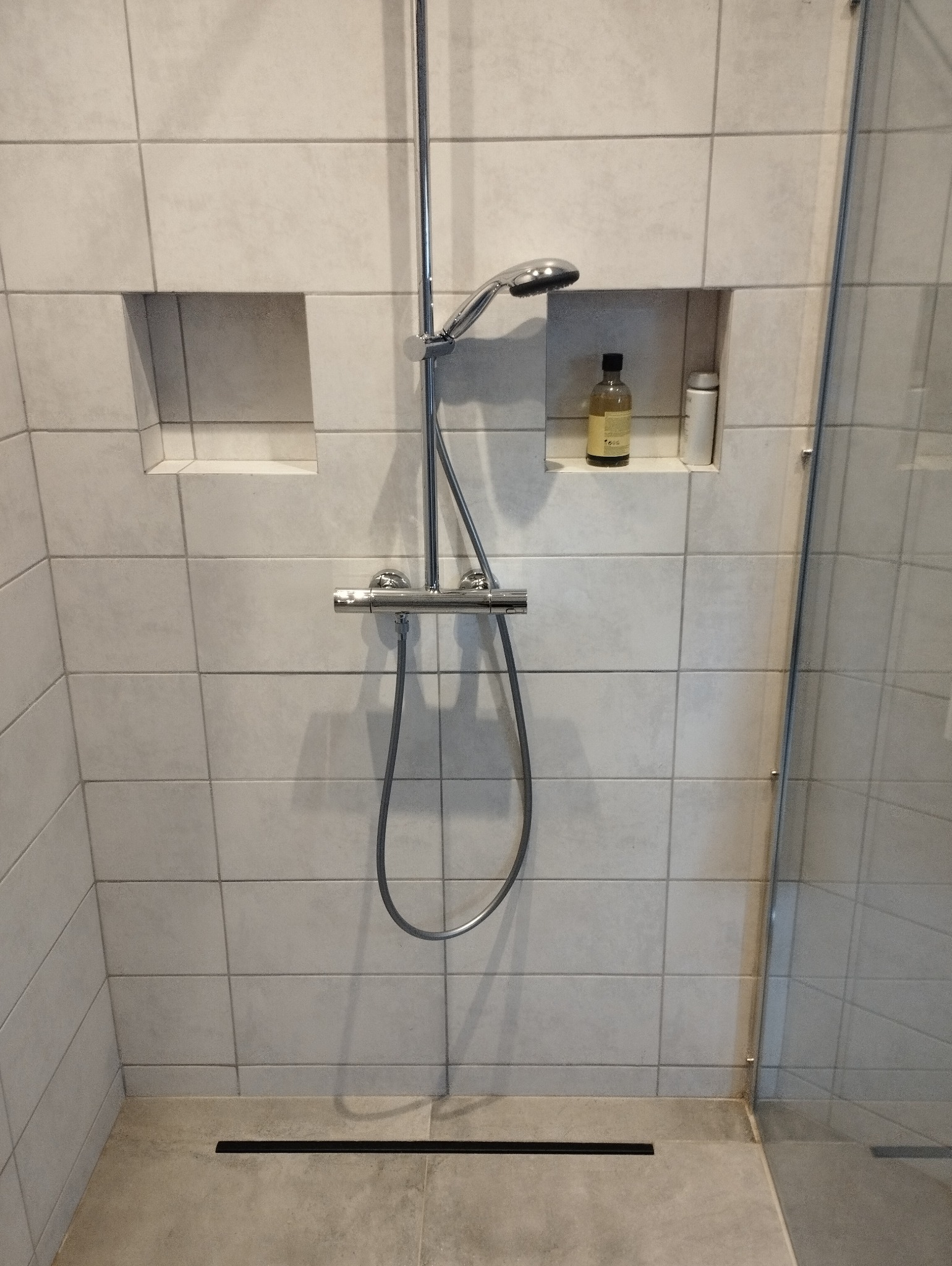 Nowoczesna kabina prysznicowa z szarymi płytkami, wbudowanymi półkami z kosmetykami, chromowaną armaturą i odpływem liniowym.