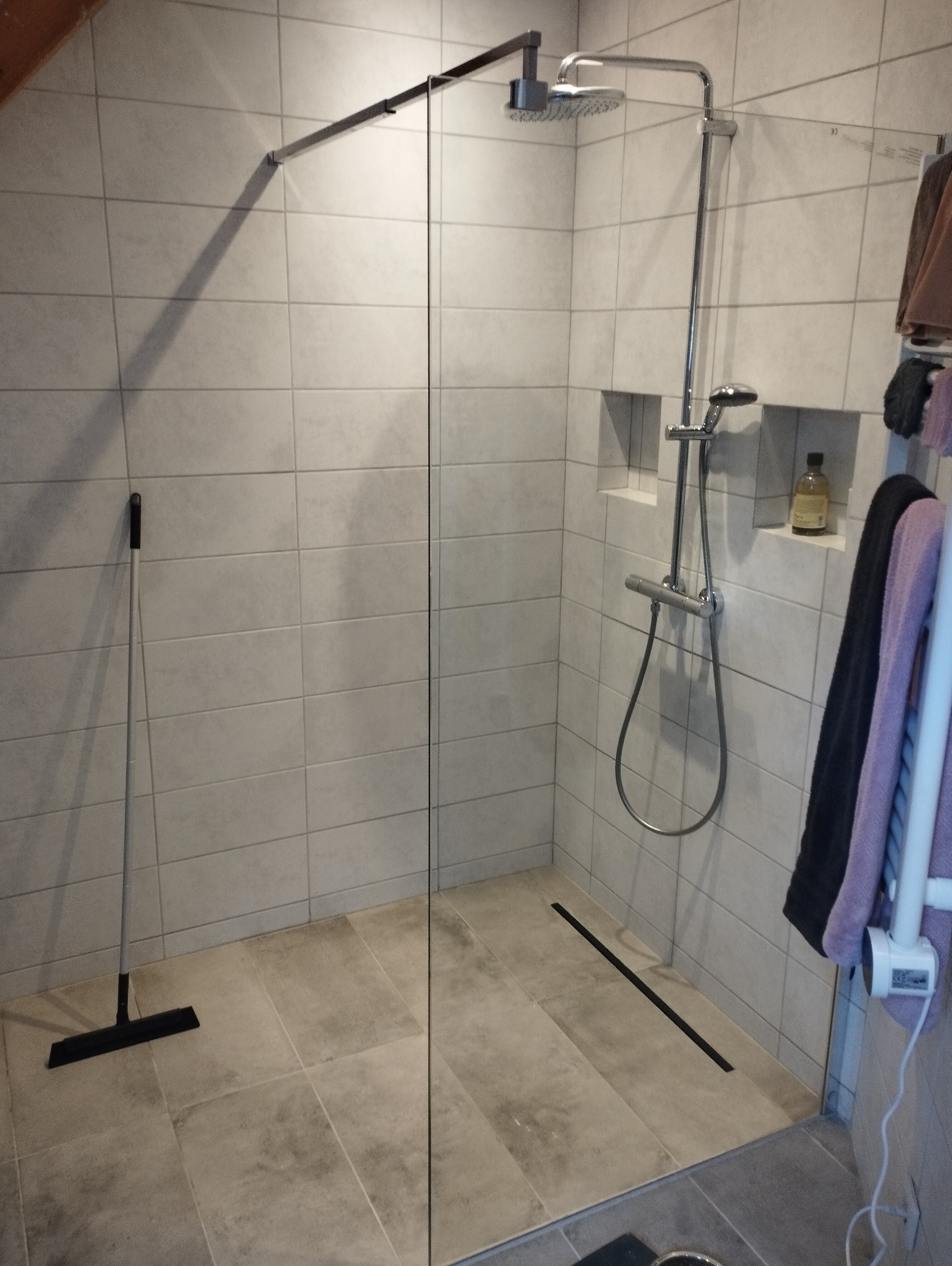 Nowoczesna kabina prysznicowa walk-in z odpływem liniowym, wykończona szarymi płytkami, z widoczną butelką kosmetyku w ściennej wnęce oraz gumową ściągaczką do wody.