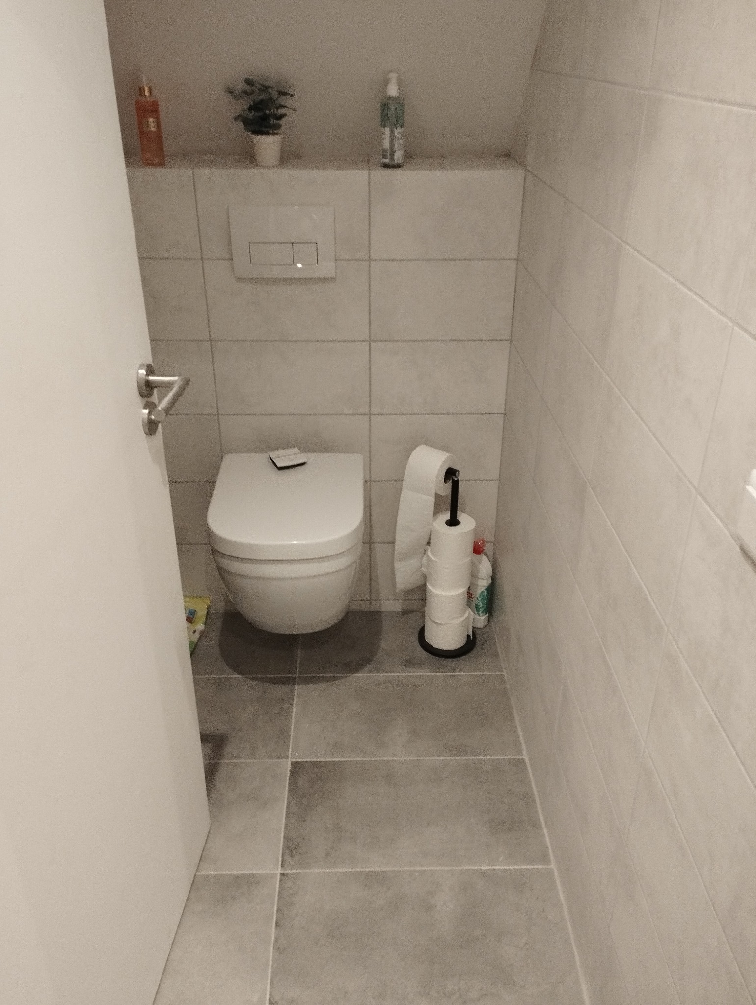 Nowoczesna toaleta z szarymi płytkami, wisząca miska WC z czarną deską, stojak na papier toaletowy obok, kosmetyki i roślina na półce nad spłuczką.