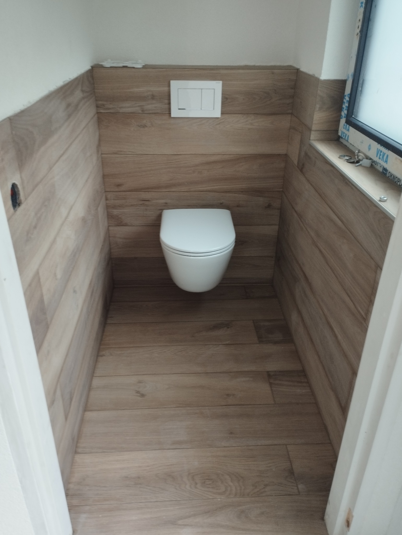 Nowoczesna toaleta podwieszana w małej łazience wykończonej płytkami imitującymi drewno, widoczne okno z profilem PCV.