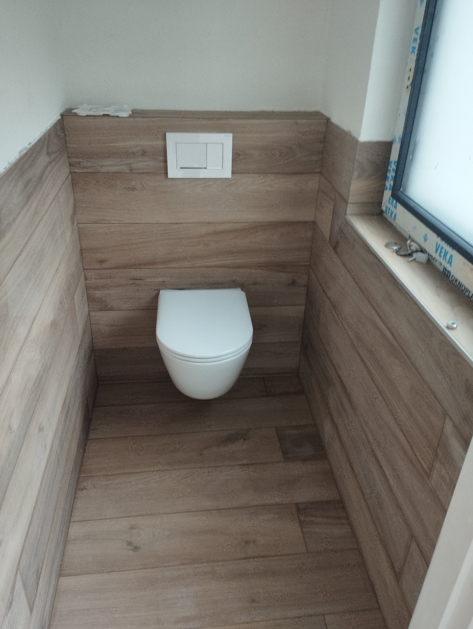 Nowoczesna toaleta podwieszana w łazience wykończonej płytkami imitującymi drewno, z widocznym przyciskiem spłukującym i oknem z profilem PCV.