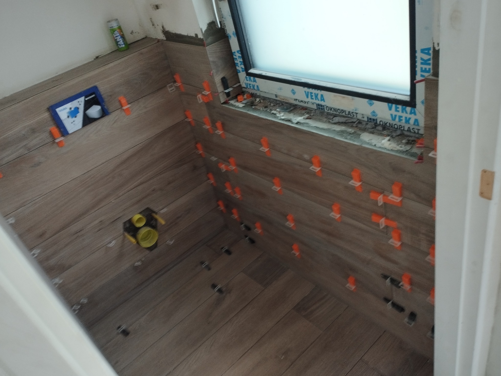 Układanie płytek imitujących drewno na ścianie i podłodze w łazience, widoczne pomarańczowe krzyżyki dystansowe, puszka z klejem i opakowanie po syfonie.